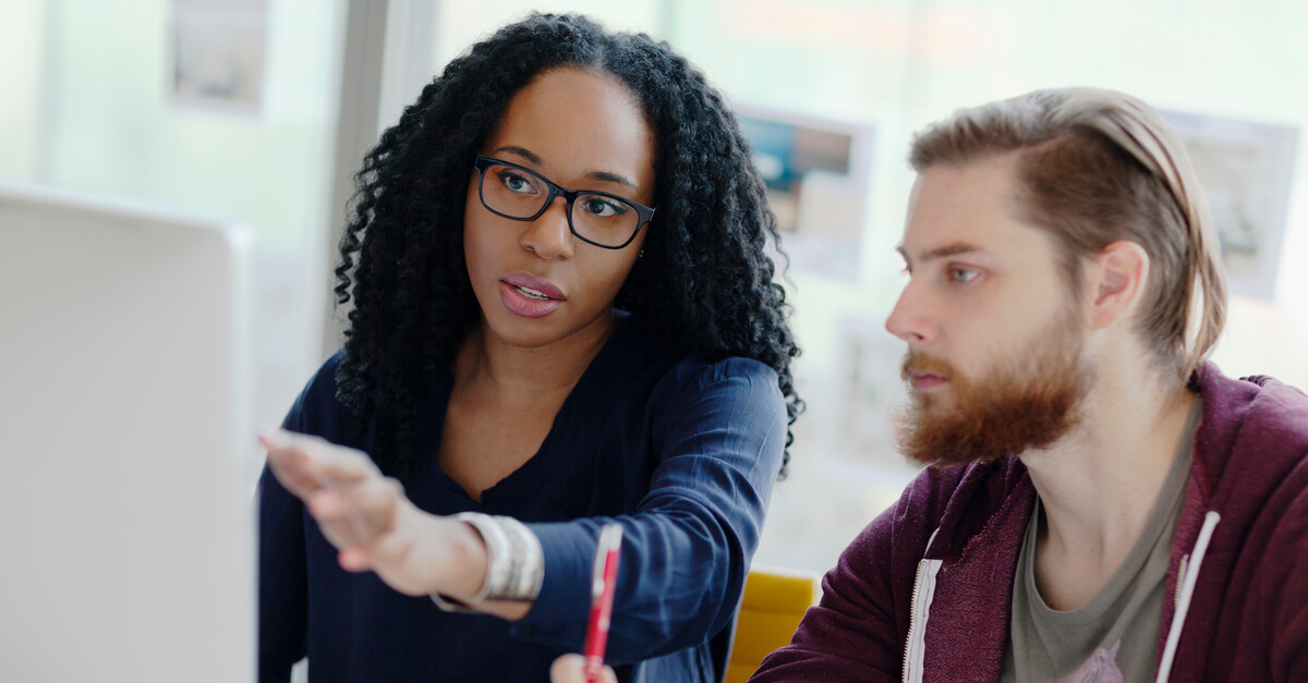 Two colleagues sit at a computer together and one points at something on the screen